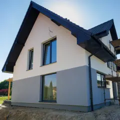 Nowy dom z dwukolorową elewacją frontową, czarnym dachem i oknami. Wokół plac budowy ze rusztowaniami.