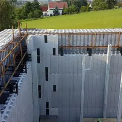 Budowa domu ze styropianu w toku. Widoczne szalunki tracone i zbrojenie. W tle zielone wzgórza i domy.