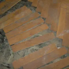 Drewniana podłoga w jodełkę, częściowo rozebrana. Widać pojedyncze deseczki, gotowe do ułożenia lub demontażu. Subit, klej do parkietu, wciąż widoczny na niektórych elementach.