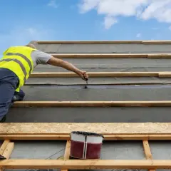 Pracownik w kamizelce zakłada papę na dach z desek, uszczelniając połączenia.