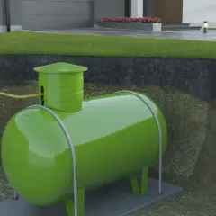 Podziemny zbiornik na gaz, prawdopodobnie izobutan, w zielonym kolorze, podłączony żółtą rurą do domu.