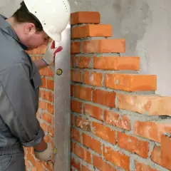 Murarz w kasku sprawdza pion muru, układając cegły. Dokładne wymiary cegły są kluczowe dla równej ściany.