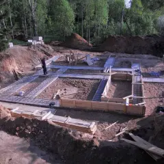 Budowa fundamentów domu. Widać zbrojenie i szalunki. Pracownicy przygotowują teren. Chudziak w tle.