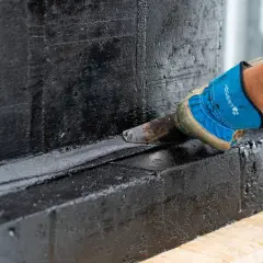 Ręka w niebieskiej rękawicy wygładza czarną masę na betonowej **fasecie**, tworząc gładką powierzchnię.