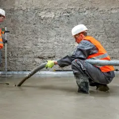 Dwóch budowlańców w kamizelkach odblaskowych wylewa pianobeton. Jeden kontroluje poziom, drugi kieruje wąż.