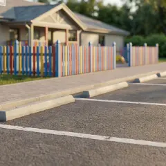 Parking spaces are visible, with concrete stops. W kolorowym ogrodzeniem i budynkiem w tle, widać odległość miejsc postojowych od granicy działki.