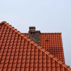 Ceglany komin na tle jasnego nieba, widok na dach pokryty czerwonymi dachówkami.
