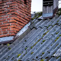 Stary dach pokryty eternitem, porośnięty mchem, z ceglanym kominem i drewnianą okapnicą.