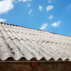 Faliste płyty eternitowe, czyli jak wygląda azbest, pokrywają dach starej ceglanej budowli. Niebo błękitne z chmurkami.