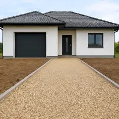 Nowy dom z białym tynkiem, czarnym dachem i bramą garażową. Szeroki podjazd z kamienia prowadzi do drzwi wejściowych.