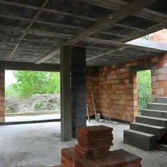 Wnętrze budowy z betonowym stropem, schodami i ceglanymi ścianami. W tle widać zielone drzewa.