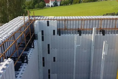 Budowa domu ze styropianu w toku. Widoczne szalunki tracone i zbrojenie. W tle zielone wzgórza i domy.