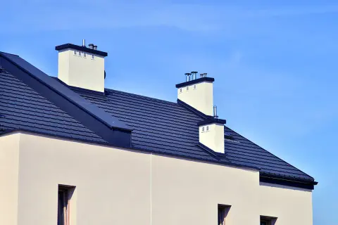 Nowoczesny dach bezokapowy z ciemnogranatową dachówką i białymi kominami, pod błękitnym niebem.