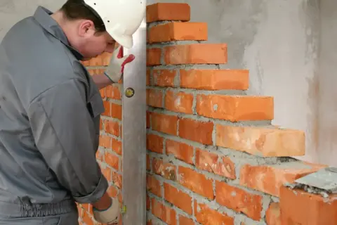 Murarz w kasku sprawdza pion muru, układając cegły. Dokładne wymiary cegły są kluczowe dla równej ściany.