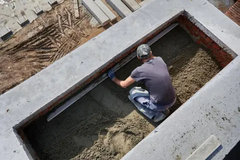 Budowlaniec wyrównuje masę, która będzie stanowiła jaki beton na fundament. Widać ceglane ściany i betonowe elementy konstrukcji.