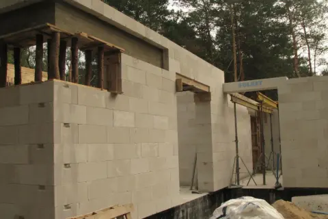 Budowa domu z bloczków betonowych. Widoczna ściana nośna z otworami okiennymi i drzwiami, podparta drewnianymi belkami.