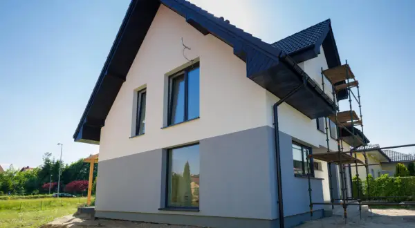 Nowy dom z dwukolorową elewacją frontową, czarnym dachem i oknami. Wokół plac budowy ze rusztowaniami.
