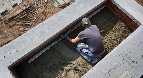 Budowlaniec wyrównuje masę, która będzie stanowiła jaki beton na fundament. Widać ceglane ściany i betonowe elementy konstrukcji.