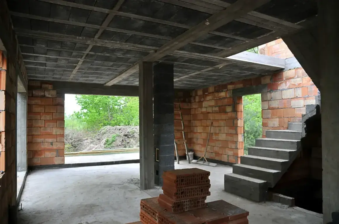 Wnętrze budowy z betonowym stropem, schodami i ceglanymi ścianami. W tle widać zielone drzewa.