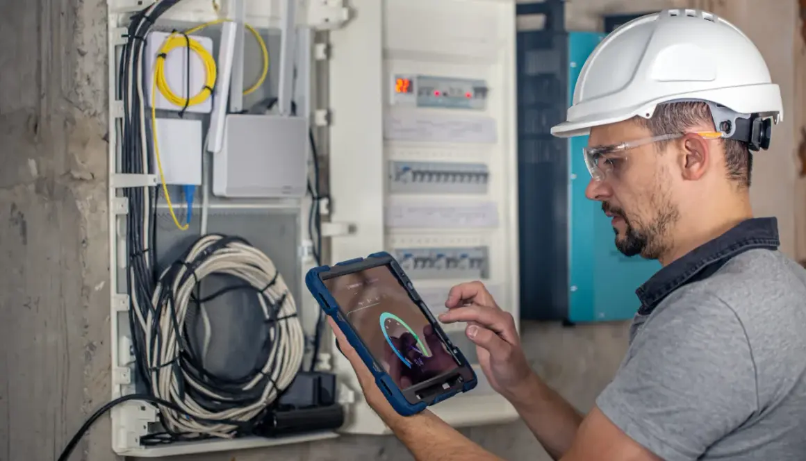 Technik w kasku i okularach ochronnych wykonuje przegląd instalacji elektrycznej, używając tabletu do monitorowania parametrów.
