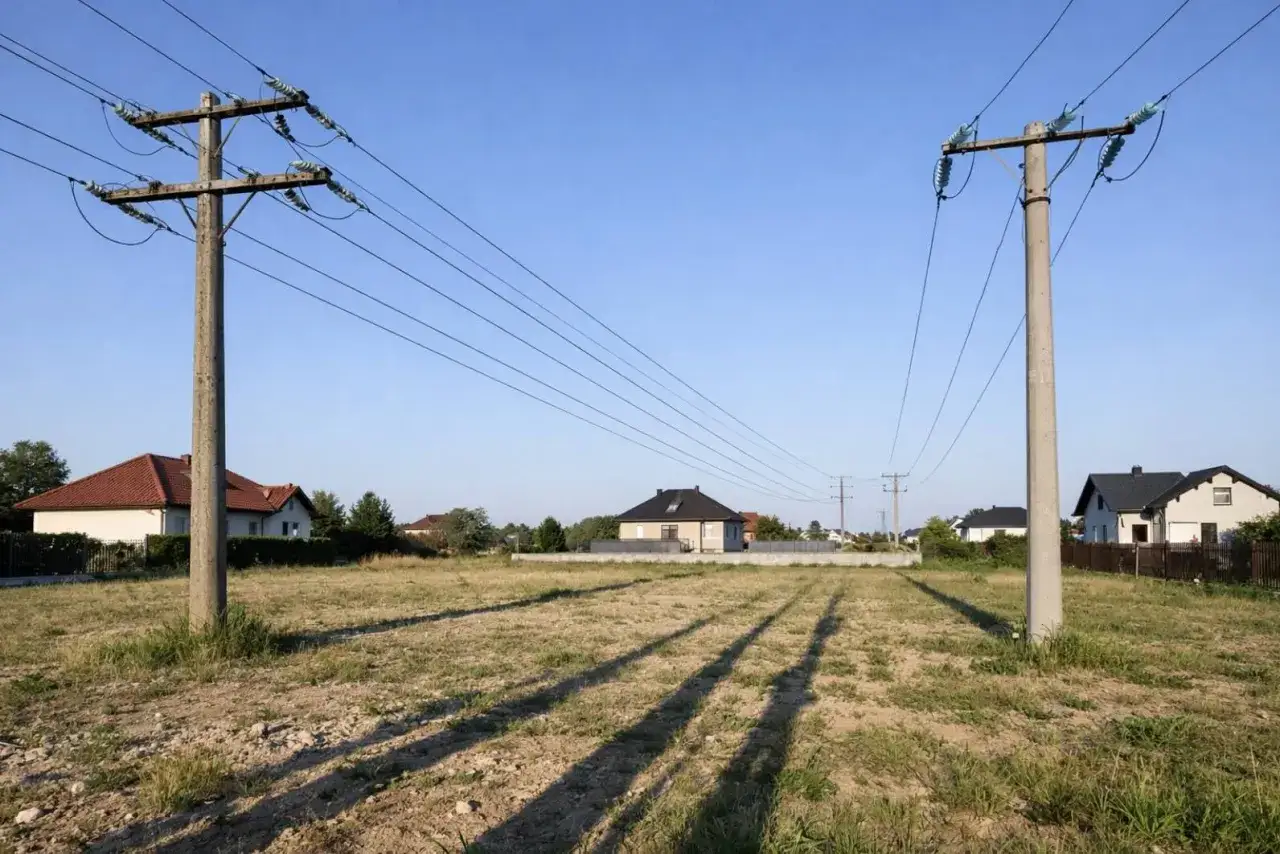 Słup energetyczny na działce, z liniami biegnącymi nad polami i domami.