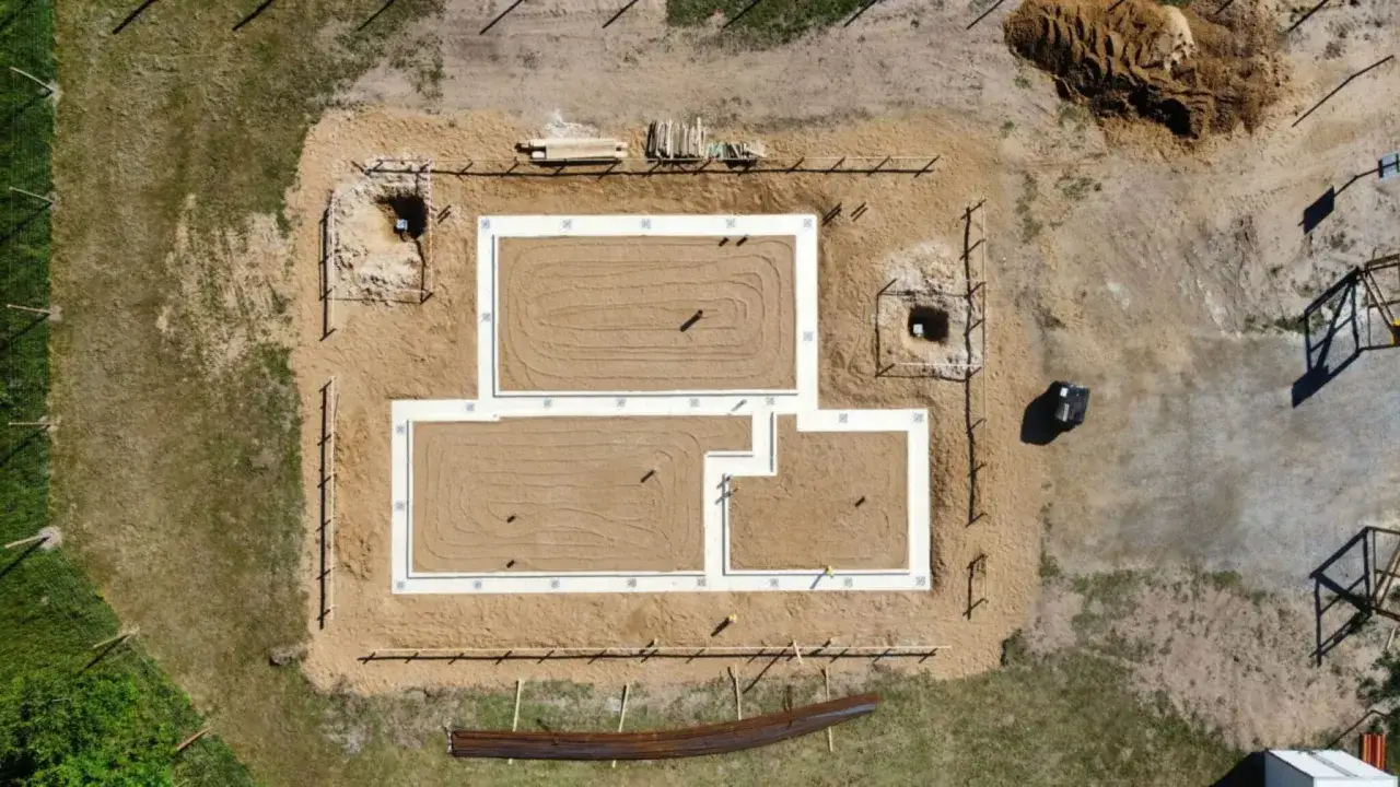 Fundament domu z chudego betonu, otoczony piaskiem i ogrodzeniem. Widać wykopy, materiały budowlane i zarys przyszłych pomieszczeń.