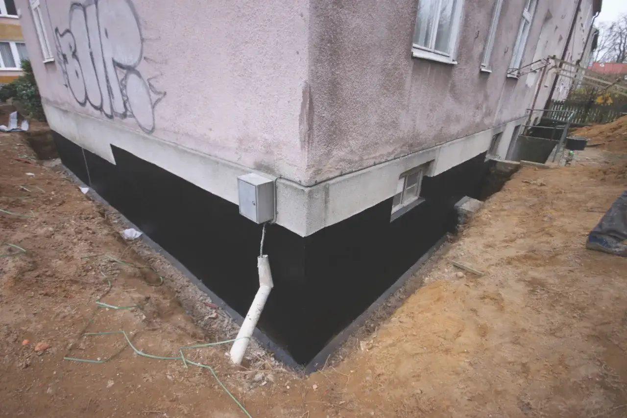 Prace przy izolacji fundamentów w starym domu. Czarna izolacja chroni ściany przed wilgocią.