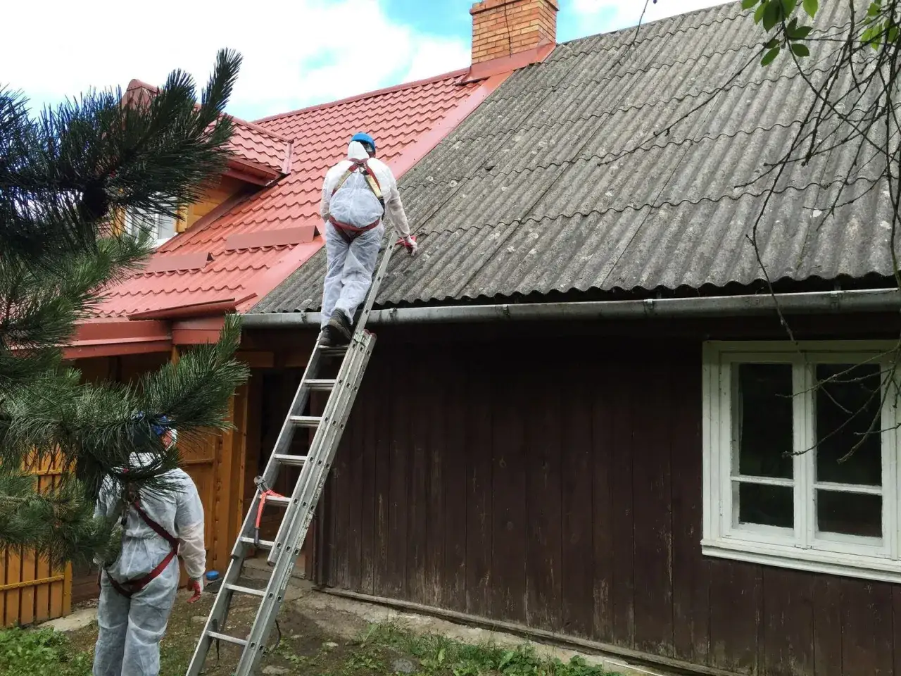 Pracownicy w kombinezonach zabezpieczających wchodzą po drabinie na dach pokryty eternitem, przygotowując się do prac.