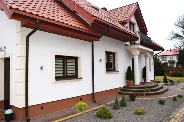 Elegancki dom z białym tynkiem i brązowym cokołem, ozdobiony kamiennymi schodami i zadbanym ogrodem.