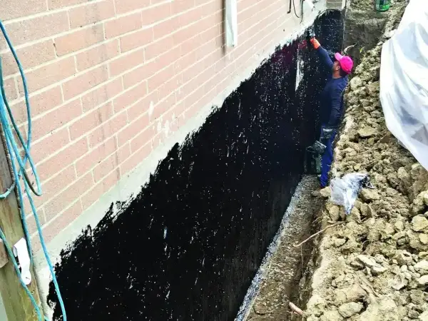 Pracownik wykonuje podbijanie fundamentów, nanosząc czarną masę izolacyjną na ścianę domu.