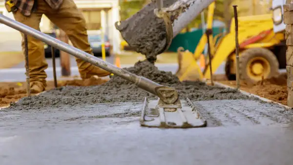 Pracownik wylewa beton na chudziaka, używając łaty do wyrównania powierzchni. W tle widać koparkę.