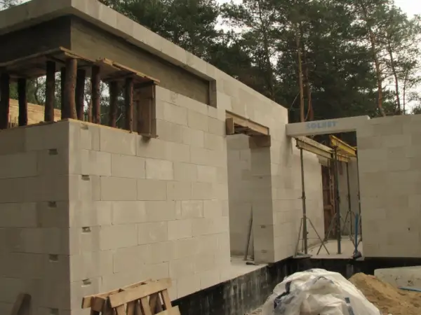 Budowa domu z bloczków betonowych. Widoczna ściana nośna z otworami okiennymi i drzwiami, podparta drewnianymi belkami.