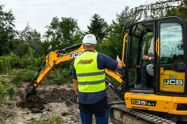 Instruktor z Akademii Operatora pokazuje, ile kosztuje godzina pracy koparki JCB. Operator w kabinie.