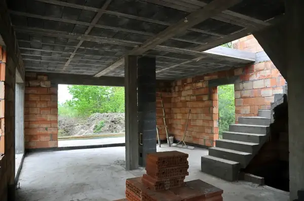 Wnętrze budowy z betonowym stropem, schodami i ceglanymi ścianami. W tle widać zielone drzewa.